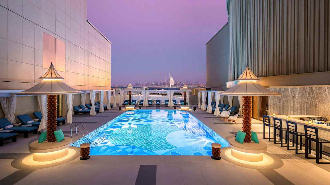 En tagterrasse med en dekorativ pool omgivet af liggestole og små parasoller. I baggrunden kan Burj Al Arab og Dubais skyline ses under en lilla aftenshimmel. (AI-genereret tekst)