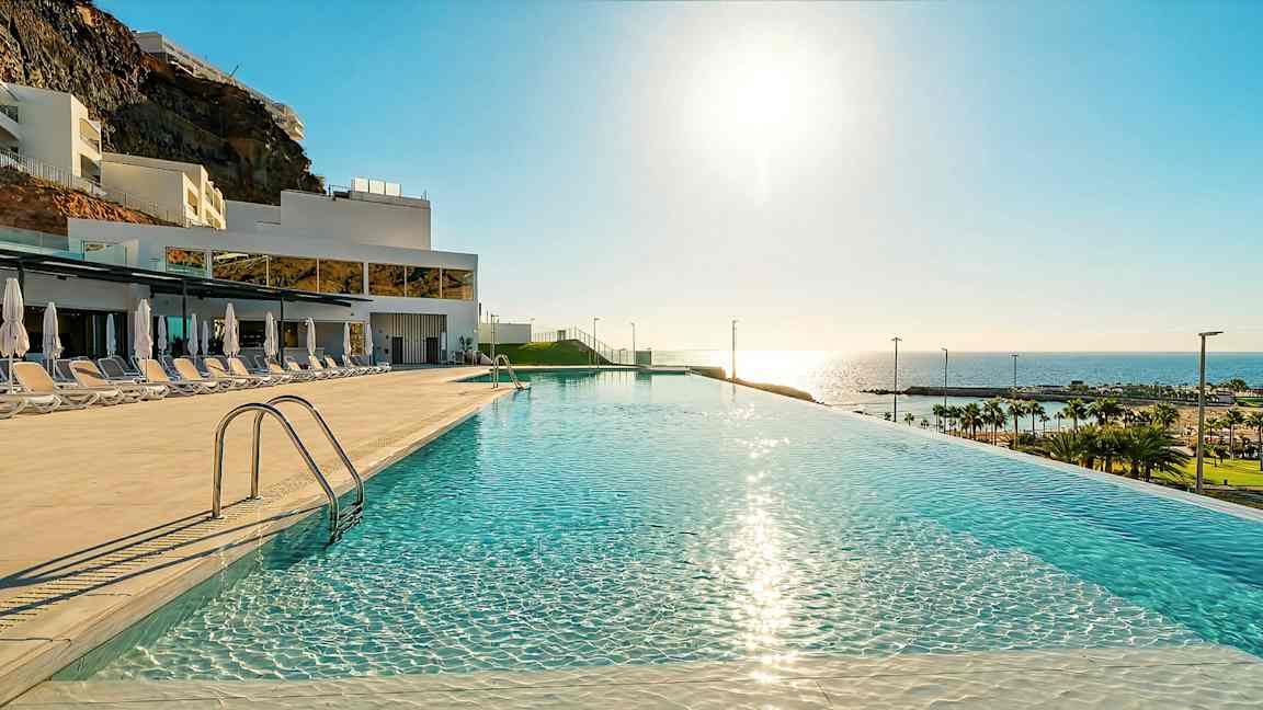 En infinitypool med spejlklart vand, solsenge og parasoller på siden, i et hotelområde med udsigt til havet og stranden. (AI-genereret tekst)
