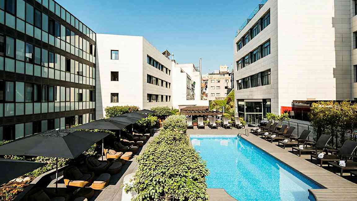 En hotelpool omgivet af liggestole og parasoller, med moderne bygninger omkring. (AI-genereret tekst)