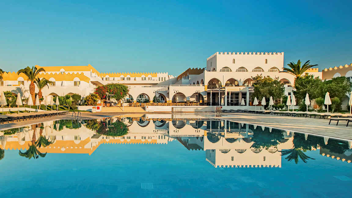 En hvid bygning med gult tag ved en pool med solsenge og parasoller. (AI-genereret tekst)