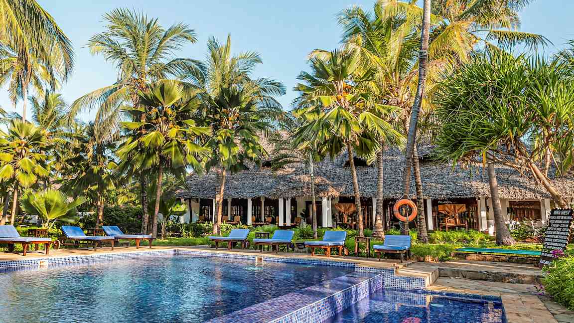 En pool omgivet af solsenge og palmer ved en bygning med stråtag. (AI-genereret tekst)