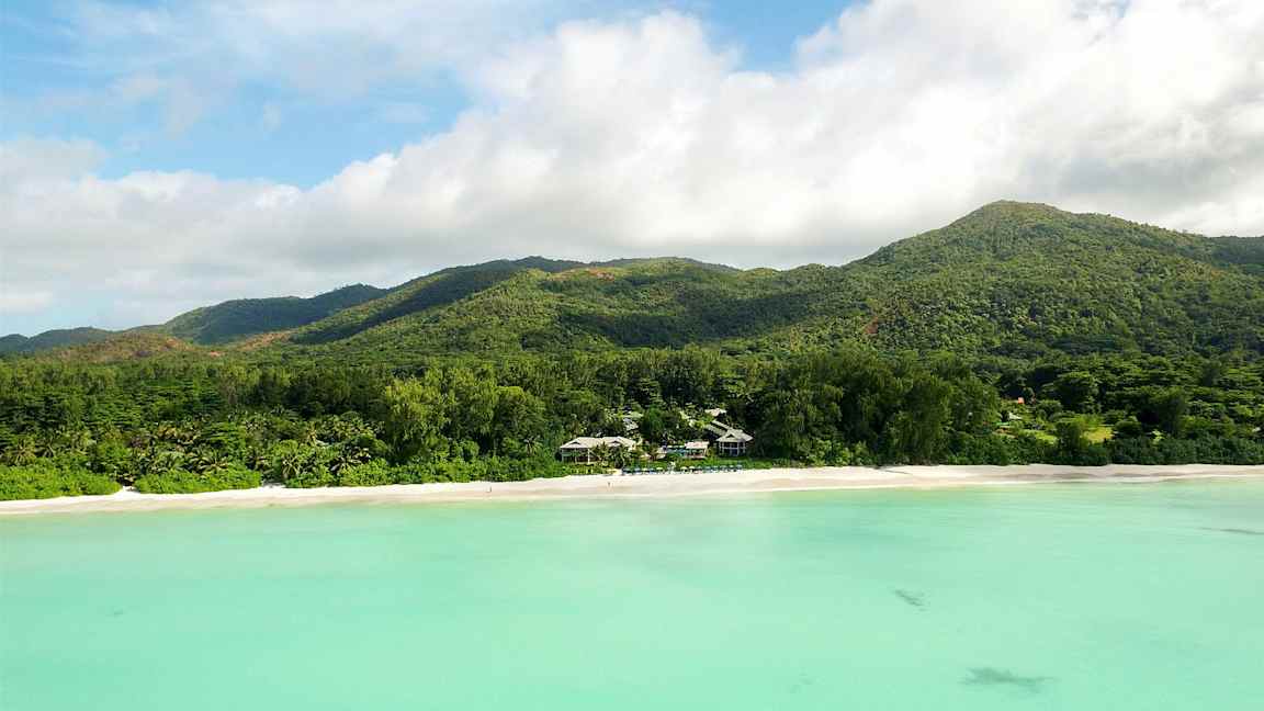 En strand med hvidt sand foran en skovklædt bjergkæde og turkisfarvet vand. (AI-genereret tekst)