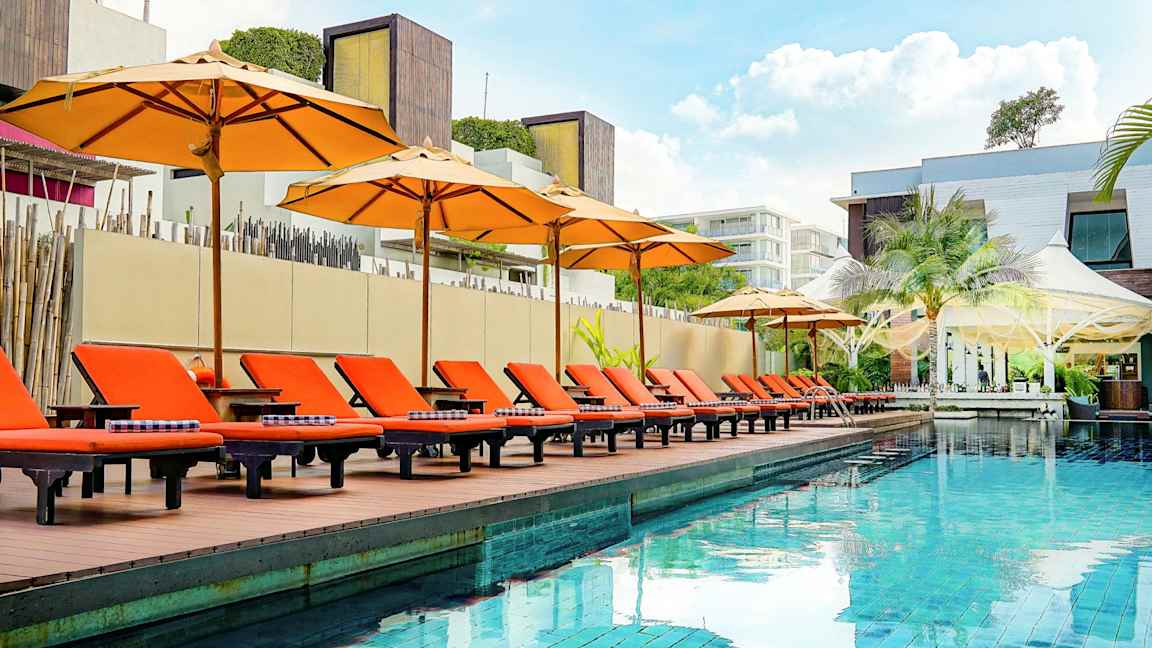 En udendørs pool med orange solsenge og parasoller ved kanten, omgivet af moderne arkitektur. (AI-genereret tekst)