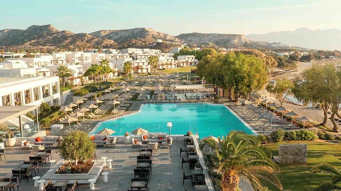 Luftfoto af et ferieanlæg med swimmingpool, liggestole, bygninger og omkringliggende landskab. (AI-genereret tekst)