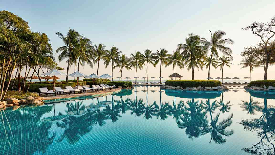 En udendørs swimmingpool med klart blåt vand, omgivet af liggestole, parasoller og palmer ved havet. (AI-genereret tekst)