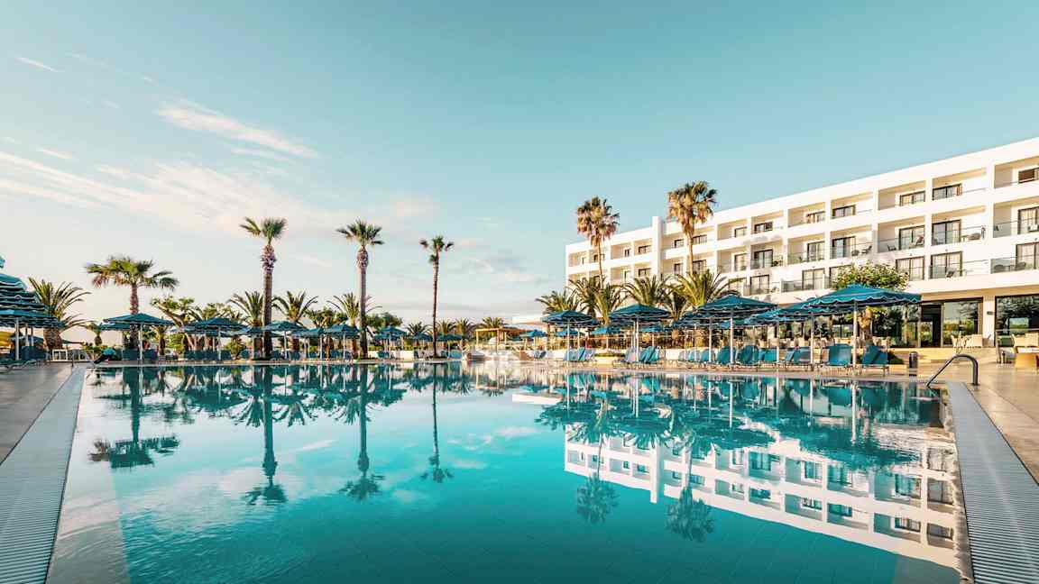 Udvendig af et hotel med en stor pool foran og omgivende palmer. (AI-genereret tekst)