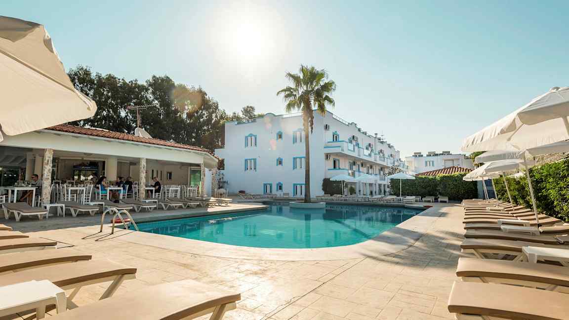 Billede af en pool med liggestole og parasoller; i baggrunden ses en hvid bygning og et åbent reservationsområde. (AI-genereret tekst)