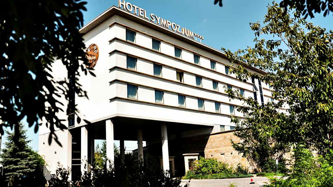 Bygning med flere etager, mærket med teksten 'Hotel Sympozjum', omgivet af træer og vegetation. (AI-genereret tekst)