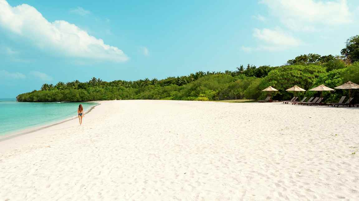 En kvinde går på en sandstrand med turkis vand og grøn vegetation i baggrunden. (AI-genereret tekst)