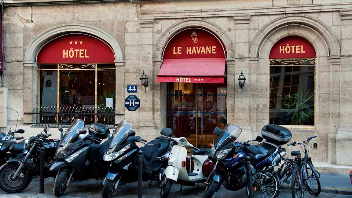 Billede, der viser indgangen til et hotel med navnet Le Havane, tre stjerner vises på markisen. Foran bygningen er der parkeret scootere og cykler. (AI-genereret tekst)