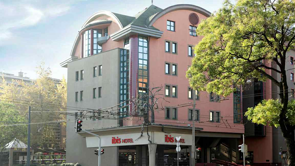 Et hotel med moderne arkitektur og flere etager, omgivet af træer og en gade med trafiklys. (AI-genereret tekst)