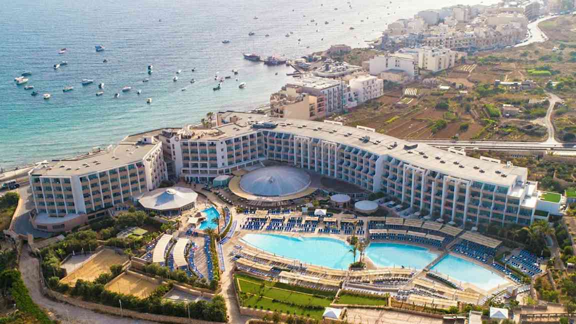 Luftfoto af et hotelkompleks med pools, grønne områder og havudsigt. (AI-genereret tekst)