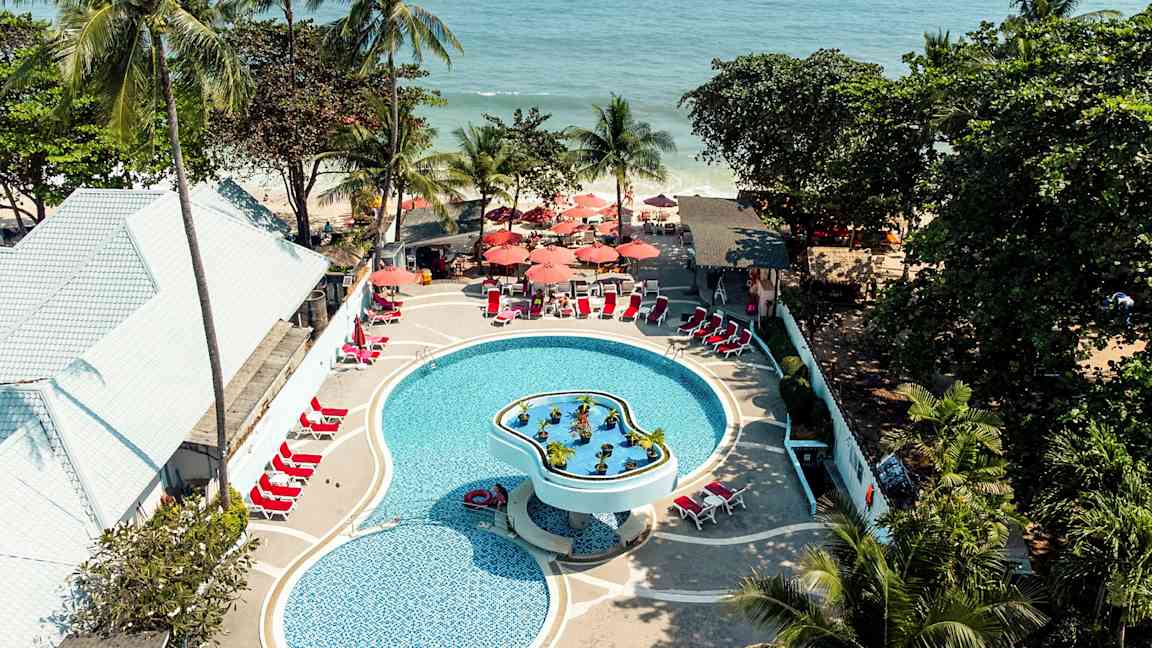 Luftfoto af en pool på et feriested med liggestole og parasoller, omgivet af tropiske træer og tæt på havet. (AI-genereret tekst)