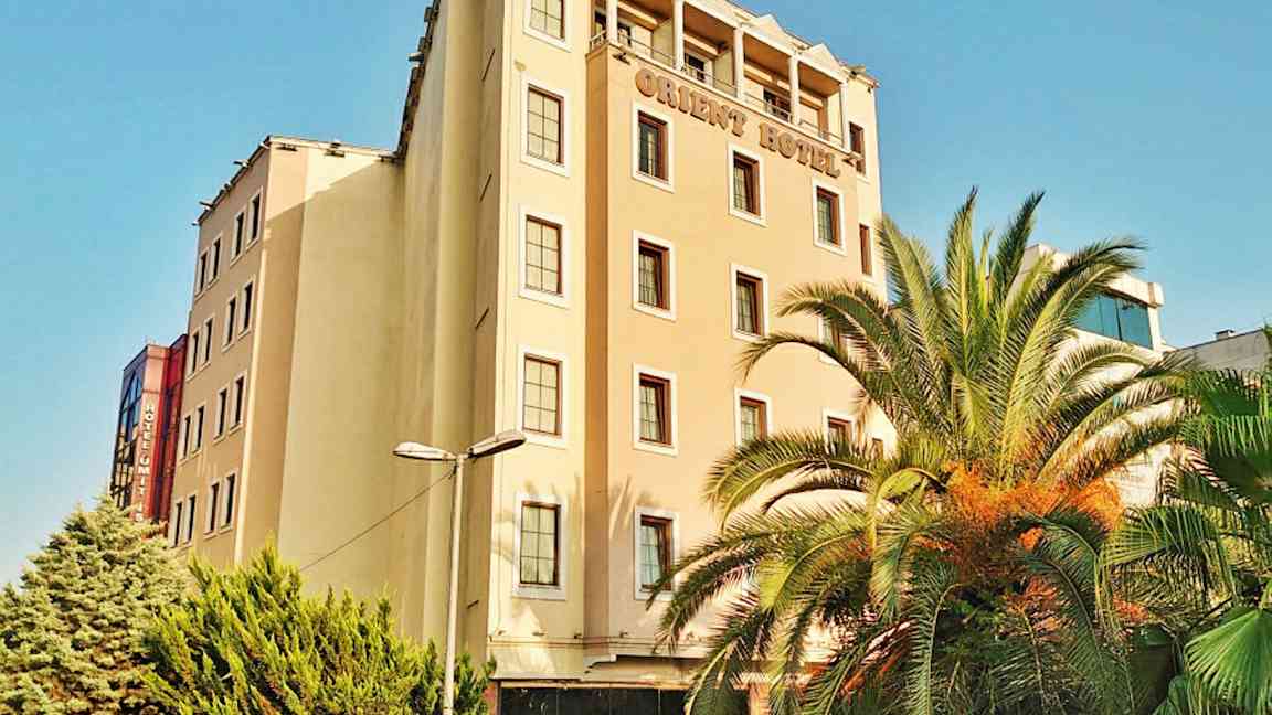 En hotelbygning med flere etager. Der er palmer og grønne træer foran bygningen. (AI-genereret tekst)