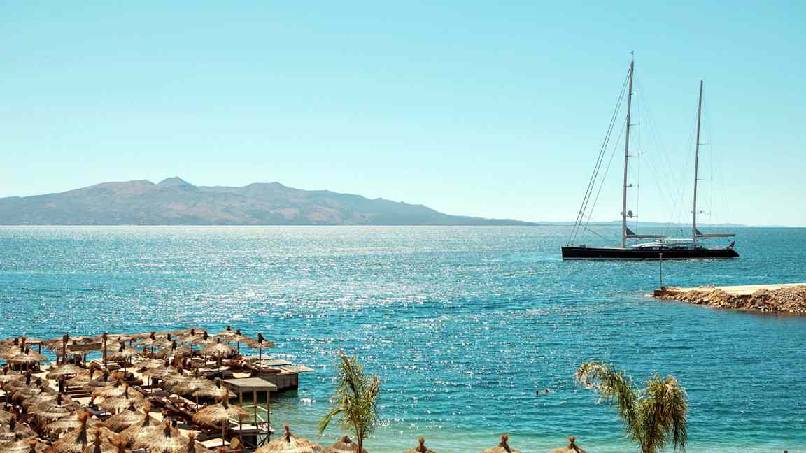 Strand med parasoller og lang sejlbåd på havet med bjerge i baggrunden. (AI-genereret tekst)