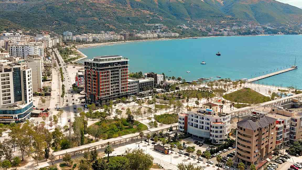 Udsigt over en kystby med bygninger, grønne områder og en kaj ved havet. (AI-genereret tekst)