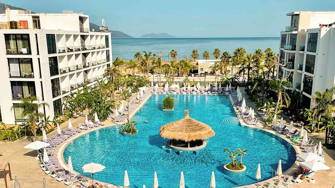 Hotelkompleks med stor swimmingpool omgivet af liggestole og parasoller, med havet i baggrunden. (AI-genereret tekst)