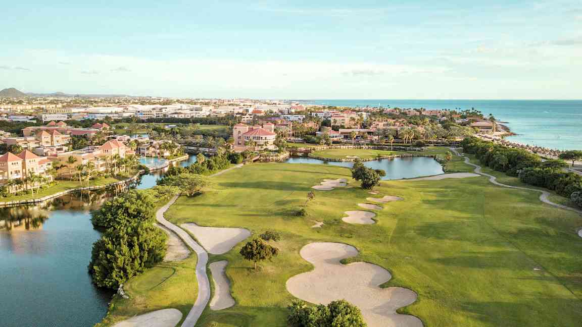 Golfbane med sandbunkere og grønne græsarealer nær vandløb og strand. Bebyggelse i baggrunden. (AI-genereret tekst)