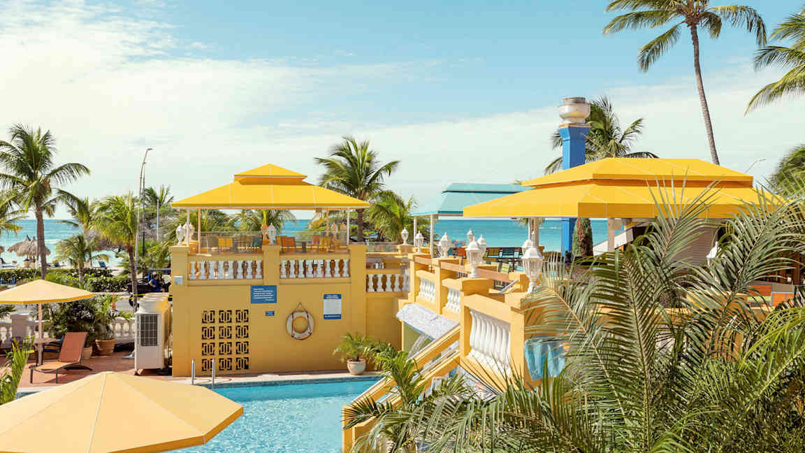 Et tropisk miljø med en swimmingpool og gule parasoller omgivet af palmer med havet i baggrunden. (AI-genereret tekst)