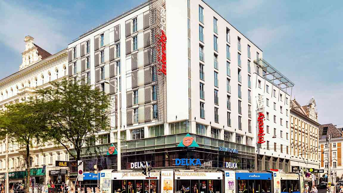 En flerfarvet sporvogn foran et moderne hotel i bylandskab. (AI-genereret tekst)