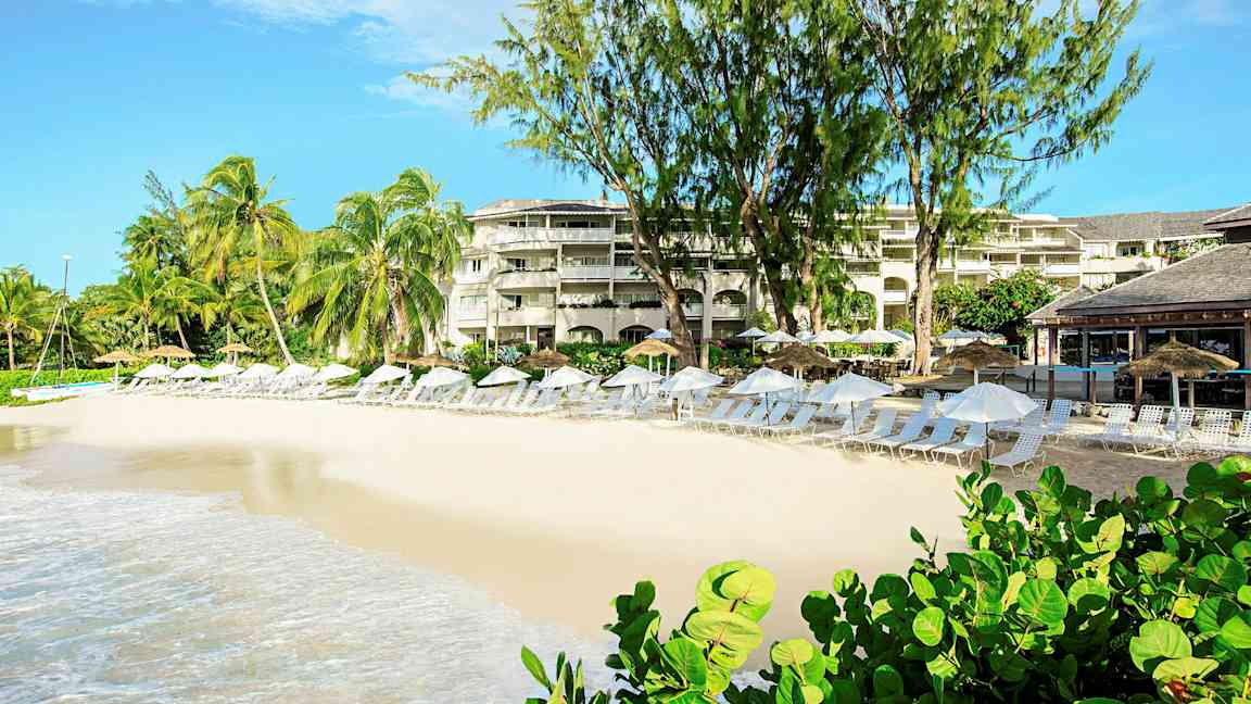 En strand med liggestole og parasoller foran en resort med palmer og beplantning. (AI-genereret tekst)