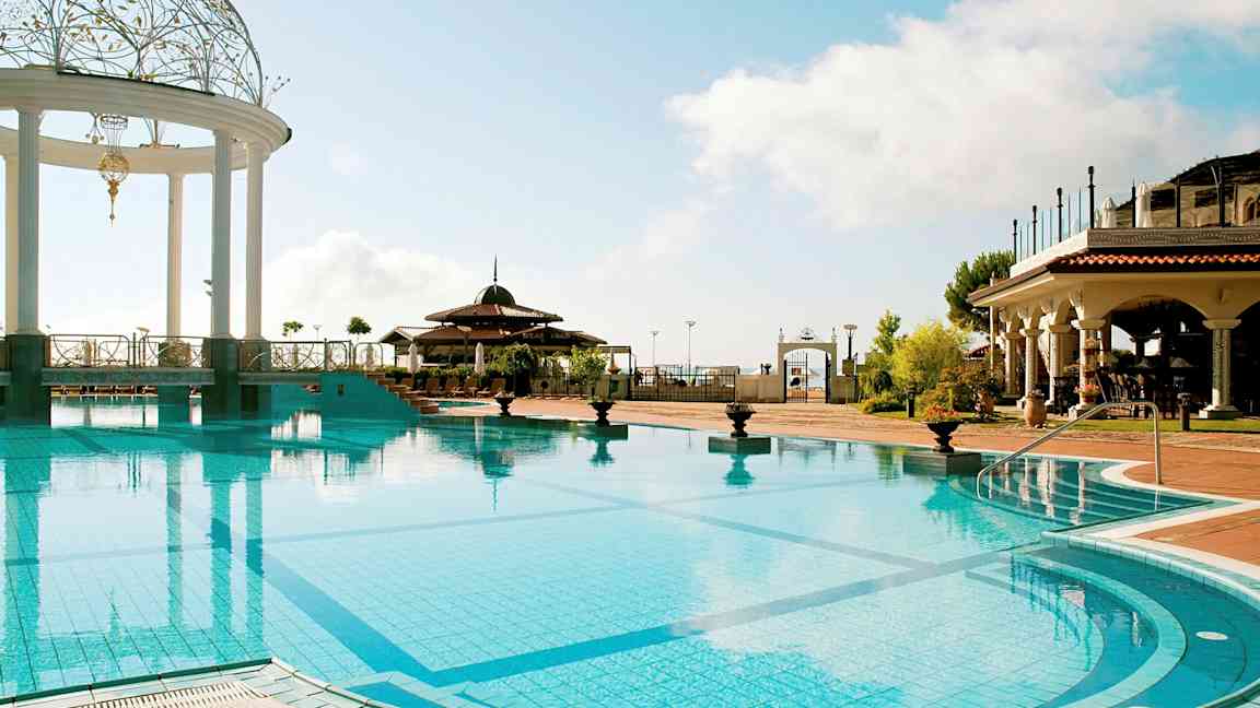 En udendørs swimmingpool med en dekoreret pavillon og omgivet af bygninger i middelhavsstil. (AI-genereret tekst)