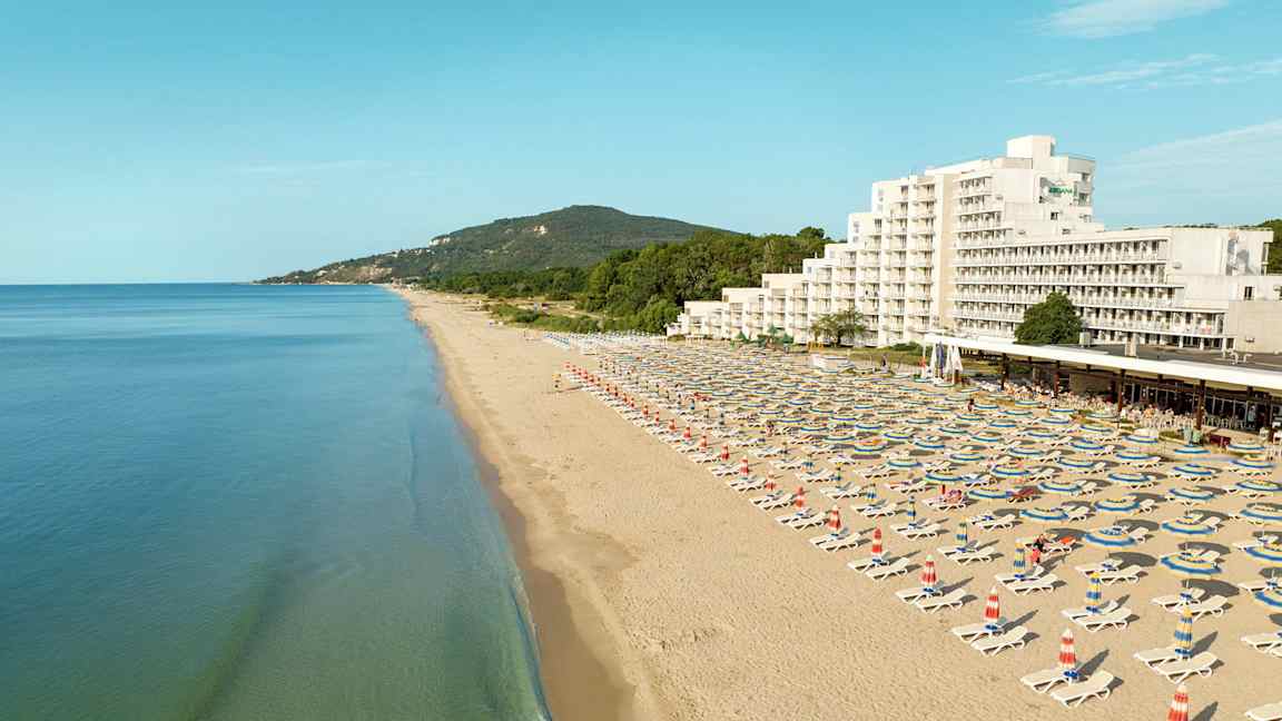 En lang sandstrand med liggestole og parasoller, samt et hotel og grønne bakker i baggrunden. (AI-genereret tekst)