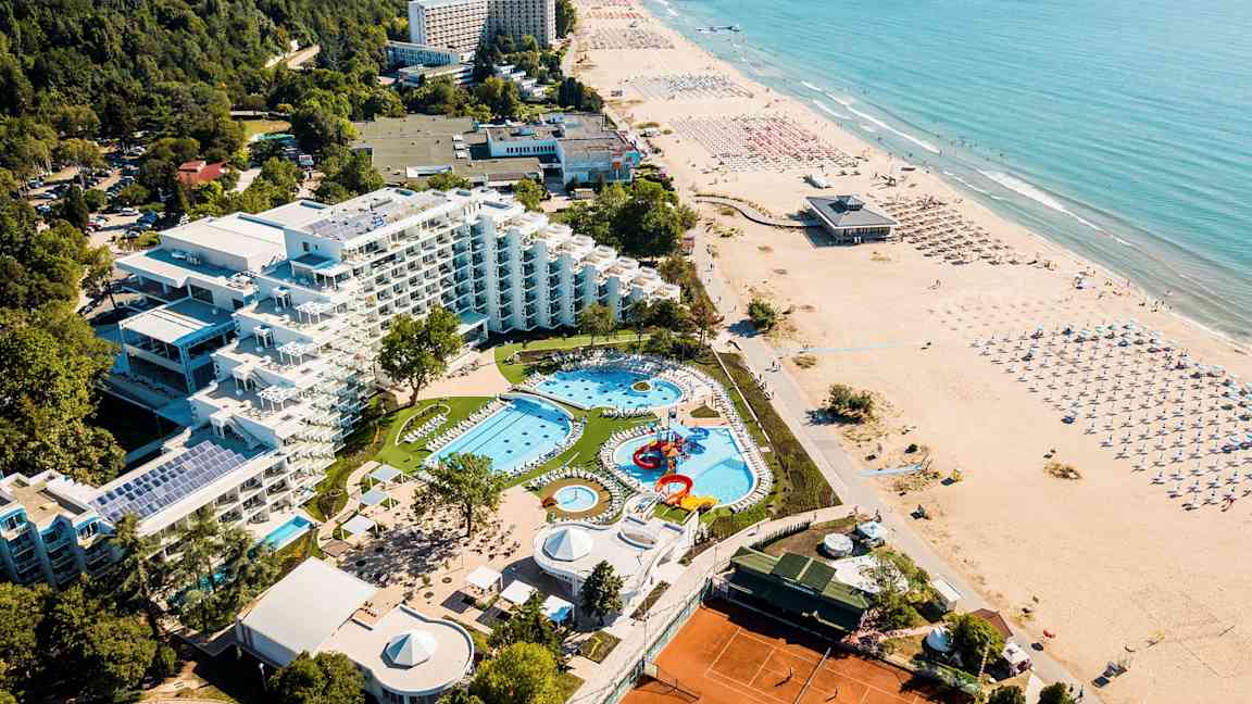 Luftfoto af hotel med poolområde, grønne omgivelser og strand. (AI-genereret tekst)