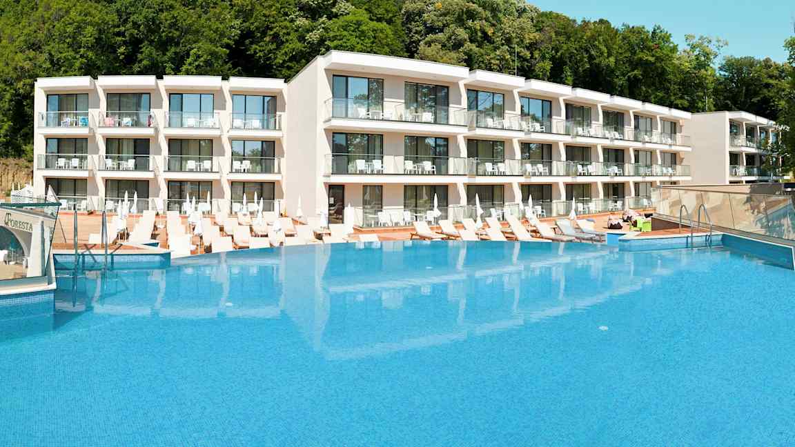 Hotelkompleks med en stor pool foran og solsenge omkring. (AI-genereret tekst)