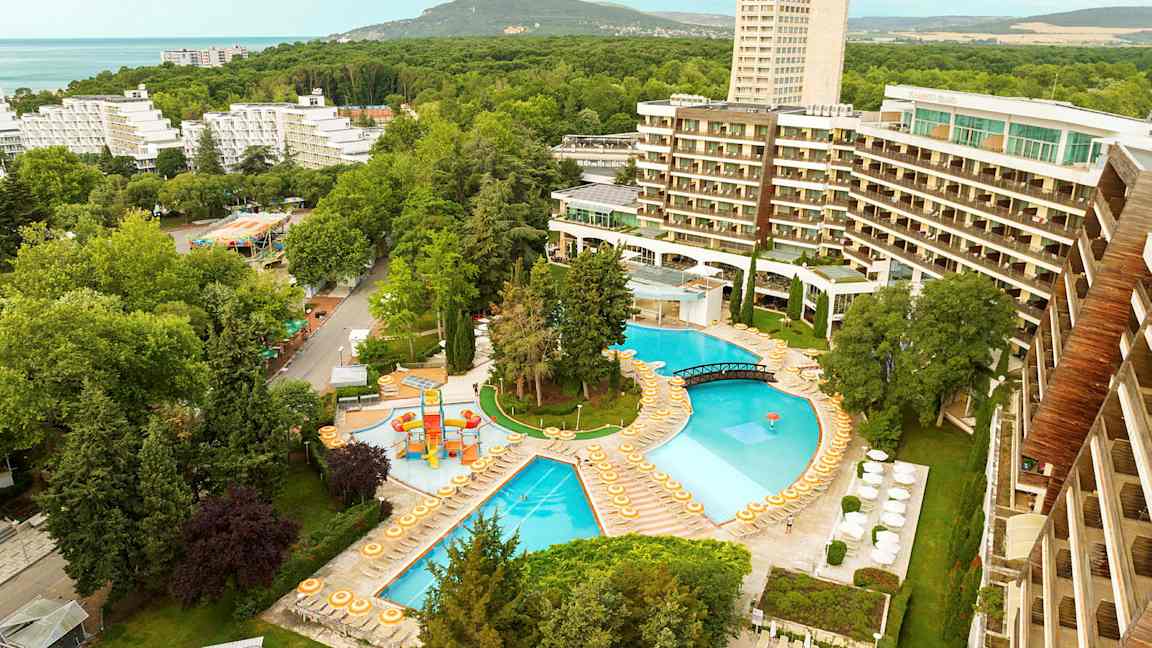 Udsigt over et hotelområde med pools, liggestole og omgivende træer. (AI-genereret tekst)