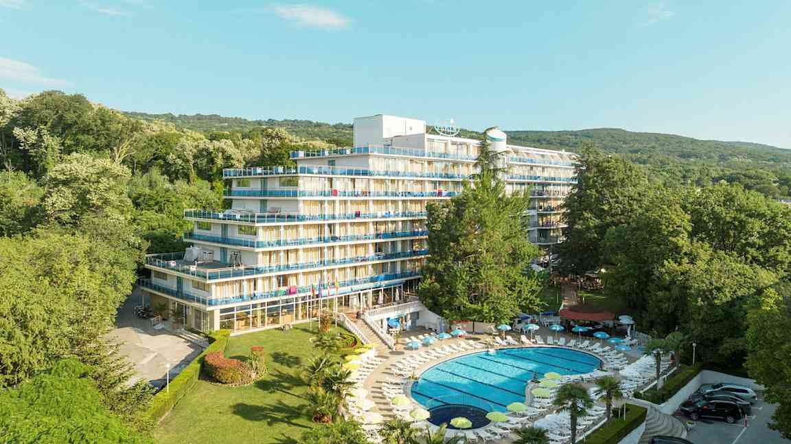 Billede af et hotel med flere etager og altaner, omgivet af frodigt grønt. En pool med liggestole ligger i forgrunden. (AI-genereret tekst)