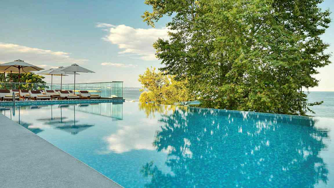 En infinitypool med liggestole og parasoller ved siden af, omgivet af træer og med udsigt over havet. (AI-genereret tekst)