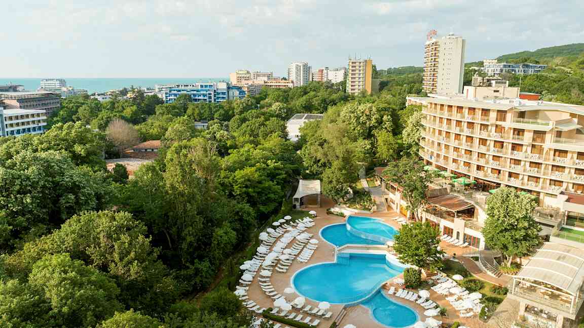 Hotelområde med pool og liggestole, omgivet af grønne områder og bygninger, med havet i baggrunden. (AI-genereret tekst)