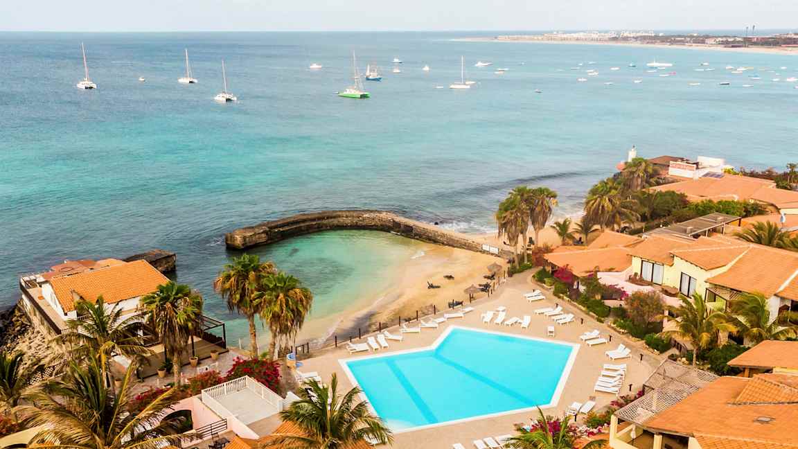 Udsigt over en badeplads og en pool ved havet, omgivet af bygninger med teglrøde tag. (AI-genereret tekst)