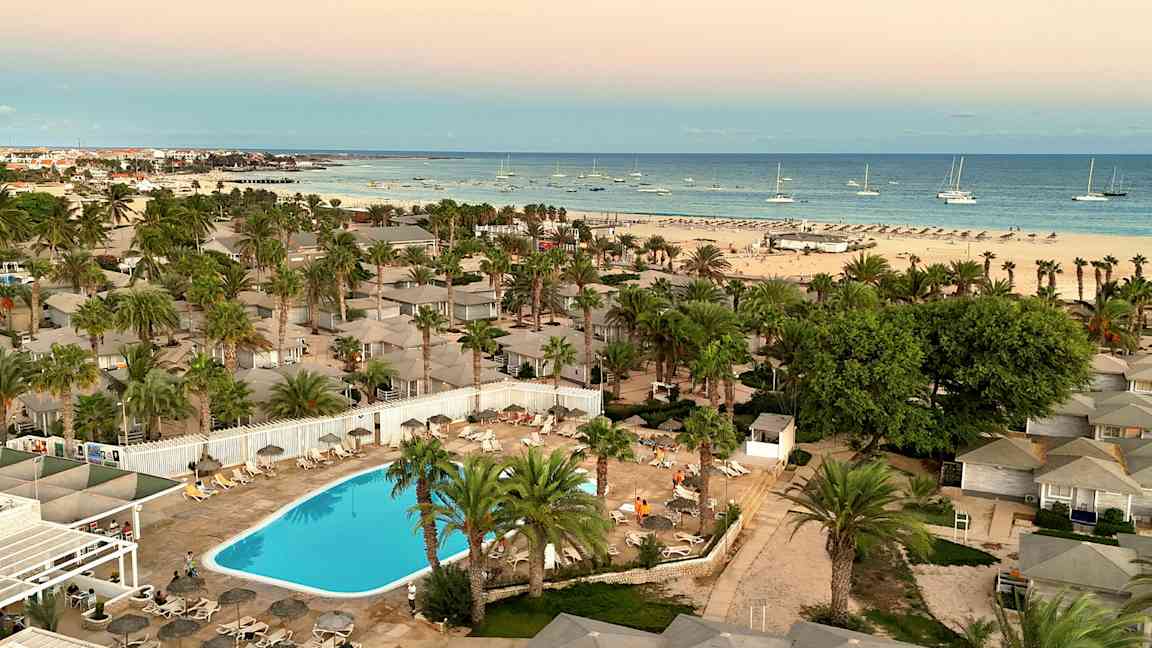 Udsigt over en resort med swimmingpool, omgivet af palmer og tæt på en strand med både på havet. (AI-genereret tekst)