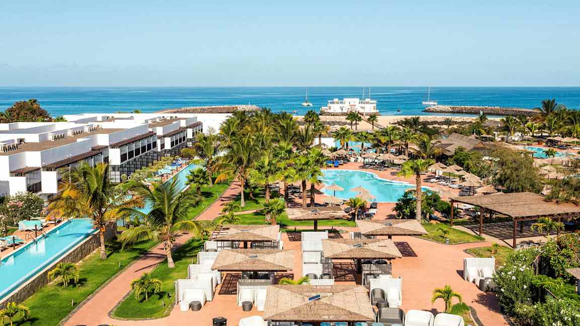 Ferieanlæg med hvide bygninger, en swimmingpool og omkringliggende grønt område, samt hav i baggrunden. (AI-genereret tekst)