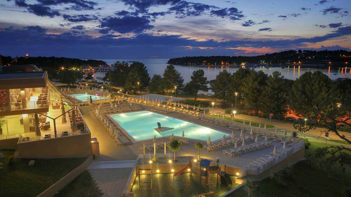 En aftenudsigt over et hotelområde med oplyste swimmingpools, omkringliggende terrasser og træer, med en sø i baggrunden. (AI-genereret tekst)
