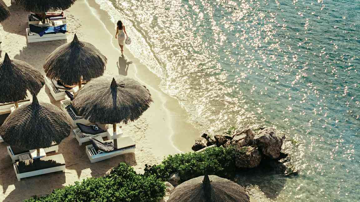 En strand med strandparasoller og liggestole, samt en person der går langs vandkanten. (AI-genereret tekst)