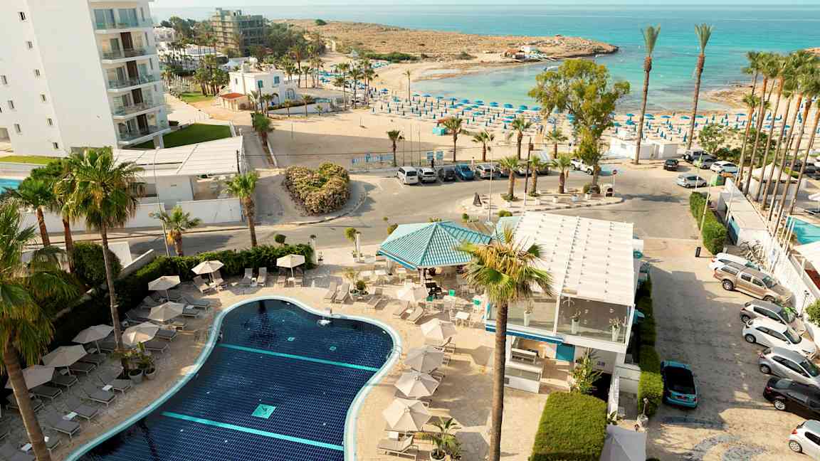 Et overblik over en hotelpool med liggestole, omgivet af palmer, med havet og en solrig strand i baggrunden. (AI-genereret tekst)