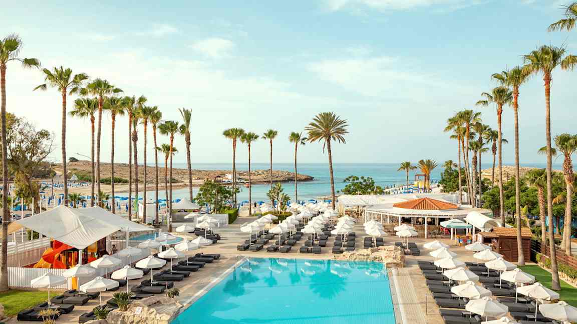 Billede af en pool med liggestole, parasoller og palmer ved en strand. (AI-genereret tekst)