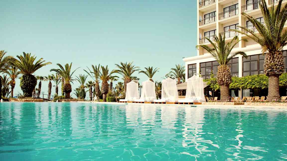 Udendørs pool med et hotel i baggrunden og omgivende palmetræer. (AI-genereret tekst)