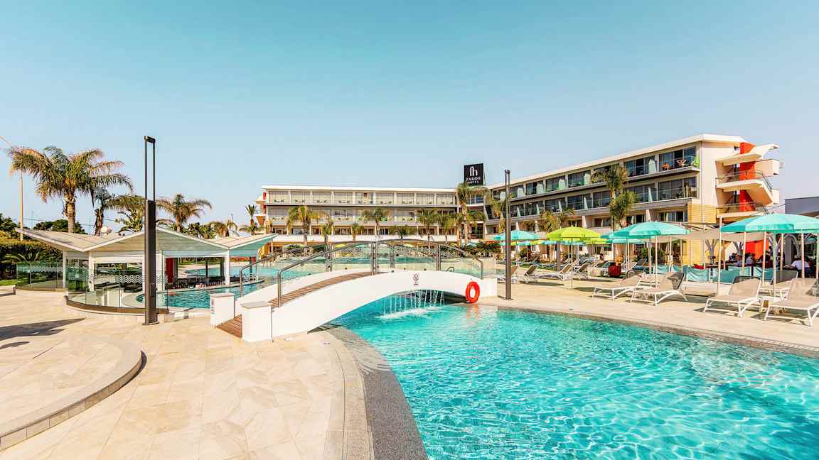 Billede af en pool med en lille bro og hotelbygning i baggrunden. (AI-genereret tekst)