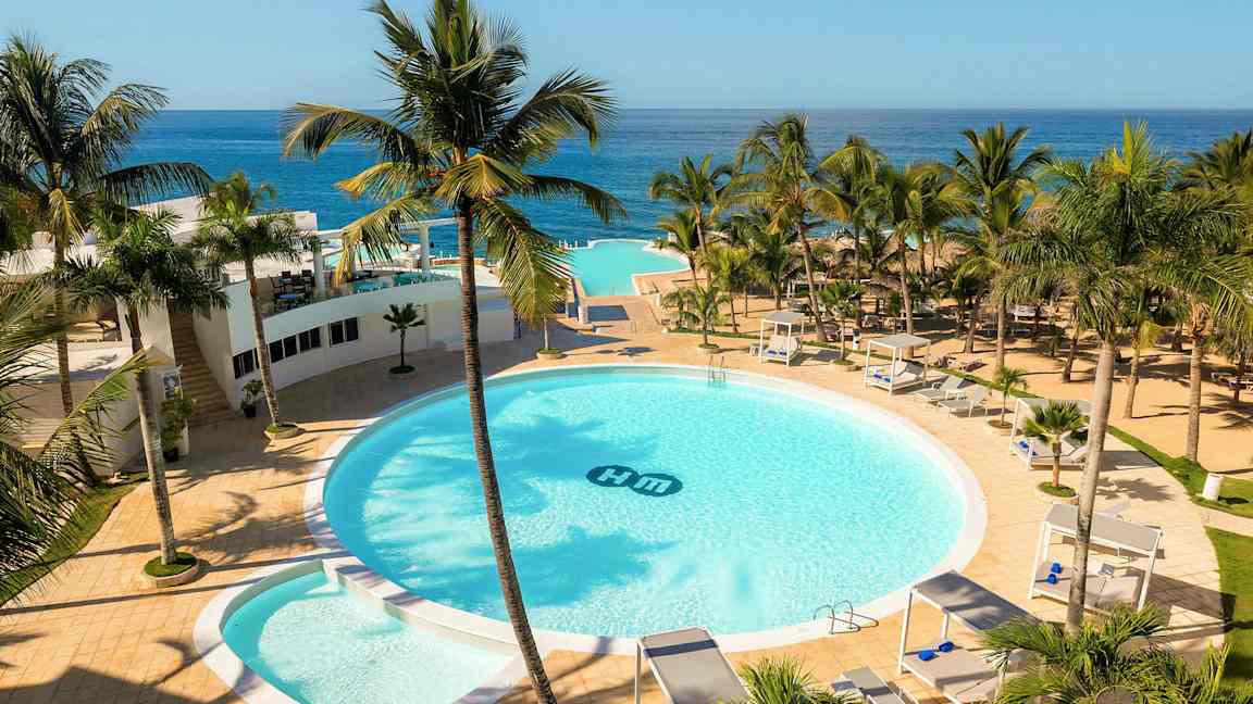Et hotelområde med en stor rund pool omgivet af palmer, solsenge og cabanaer. Havudsigt i baggrunden. (AI-genereret tekst)