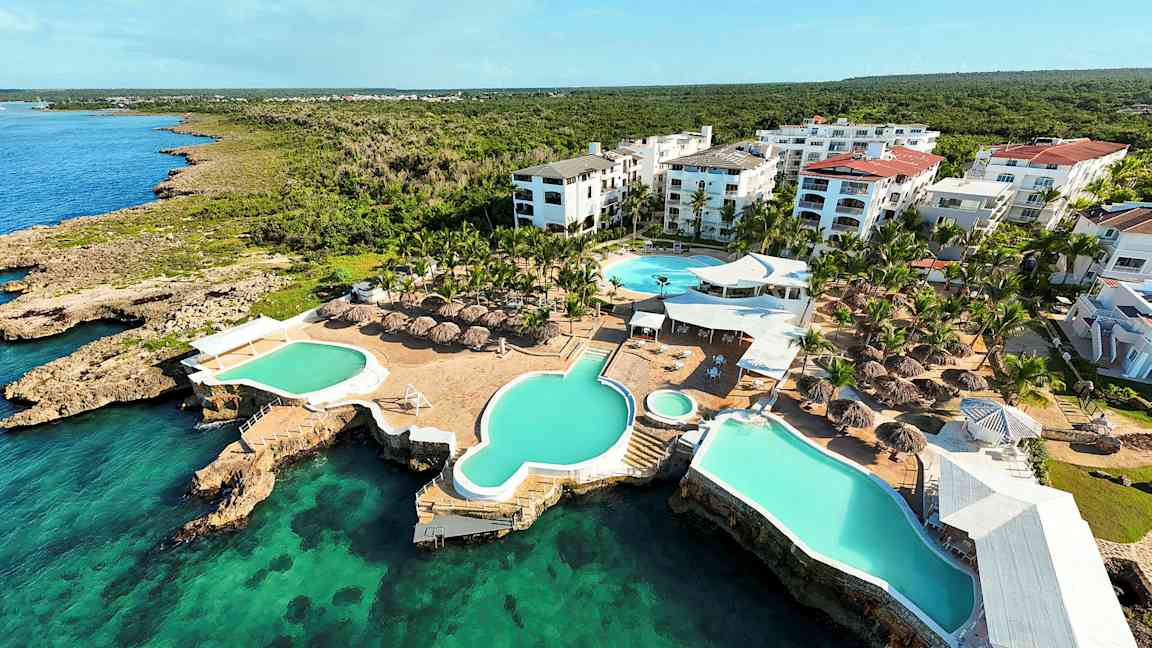 Luftfoto af en ferieanlæg med swimmingpools og baldakiner ved havet. (AI-genereret tekst)