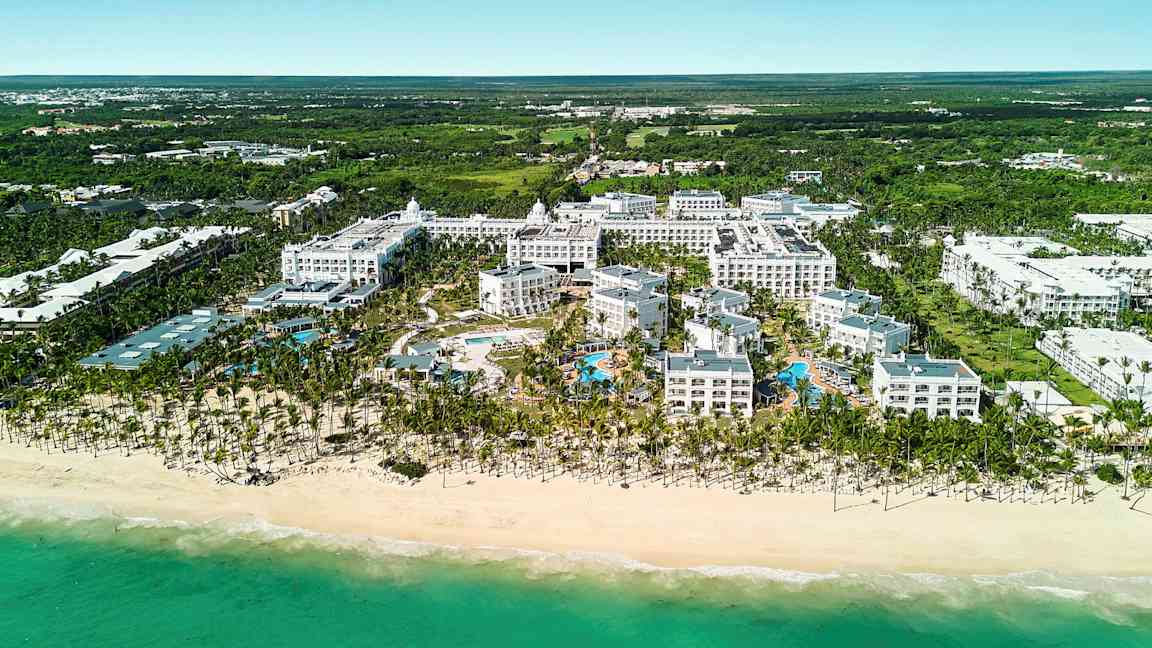 Luftfoto af et hotelkompleks ved en strand, inklusive poolområde, omgivet af grønne omgivelser. (AI-genereret tekst)