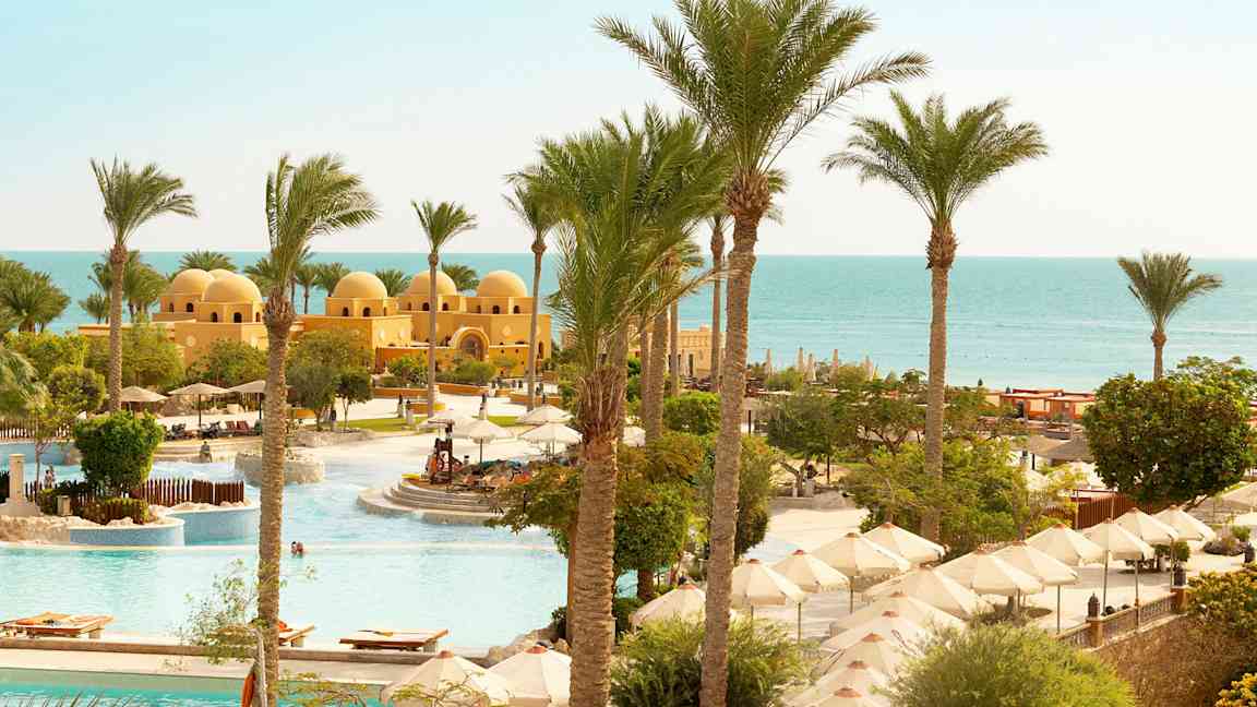 Udsigt over et poolområde med parasoller, palmer og en gul bygning med kupler tæt på havet. (AI-genereret tekst)