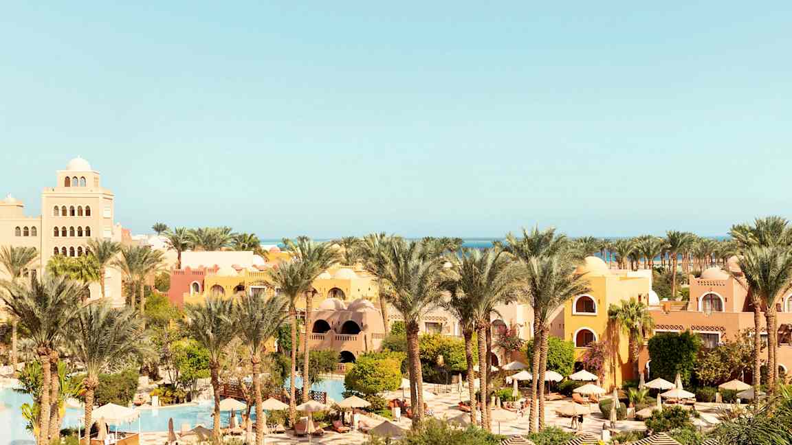 Et feriested med gule og orange bygninger, omgivet af palmer og grønt, med swimmingpools og parasoller. (AI-genereret tekst)