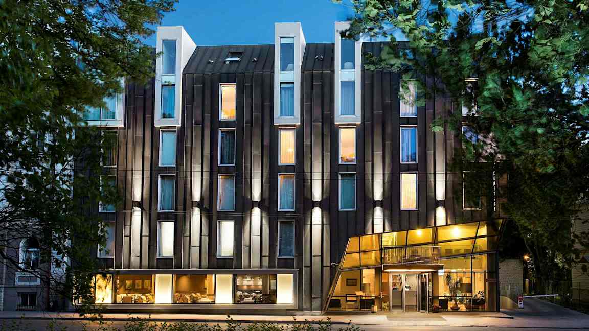 Moderne hotels facade med glas og oplyste vinduer omgivet af træer. (AI-genereret tekst)