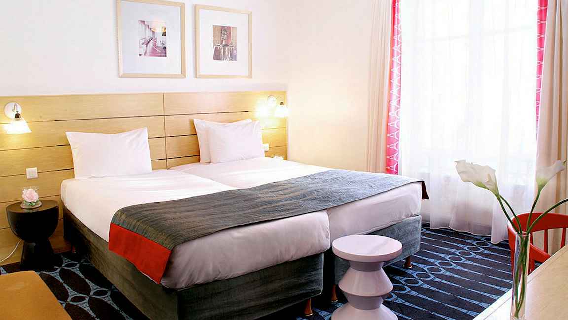 Et hotelværelse med to enkeltsenge, et dekorativt sengegærde og et vindue med lysindfald. (AI-genereret tekst)