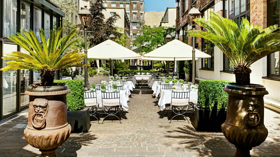 Udendørs selskabsområde med borde og parasoller omgivet af bygninger og palmer. (AI-genereret tekst)